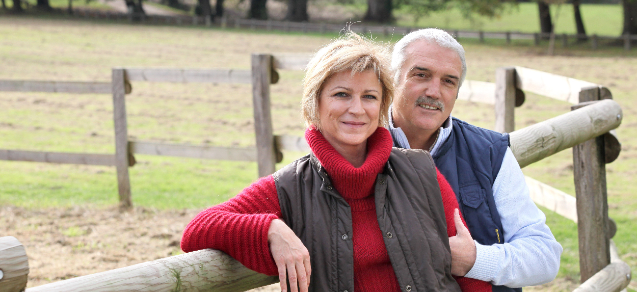 An older couple leaning against a farm fence