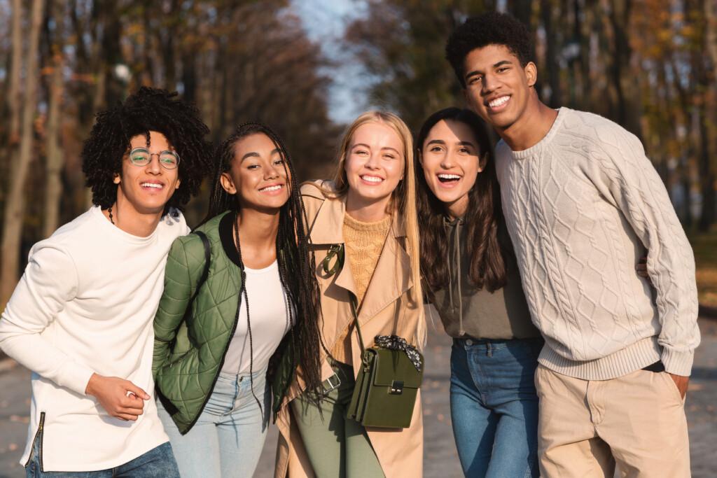 A group of five smiling teenagers standing outdoors with their arms around each others' shoulders