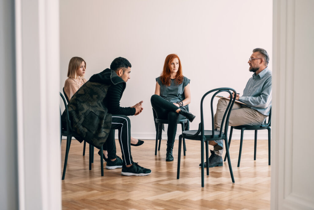 Group therapy session with a man with clipboard and three young people sitting in a circle of chairs