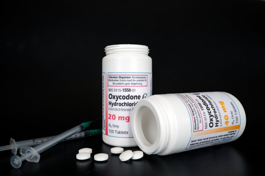 Oxycodone Hydrochloride prescription bottles in background with pills and syringes laying in front. 