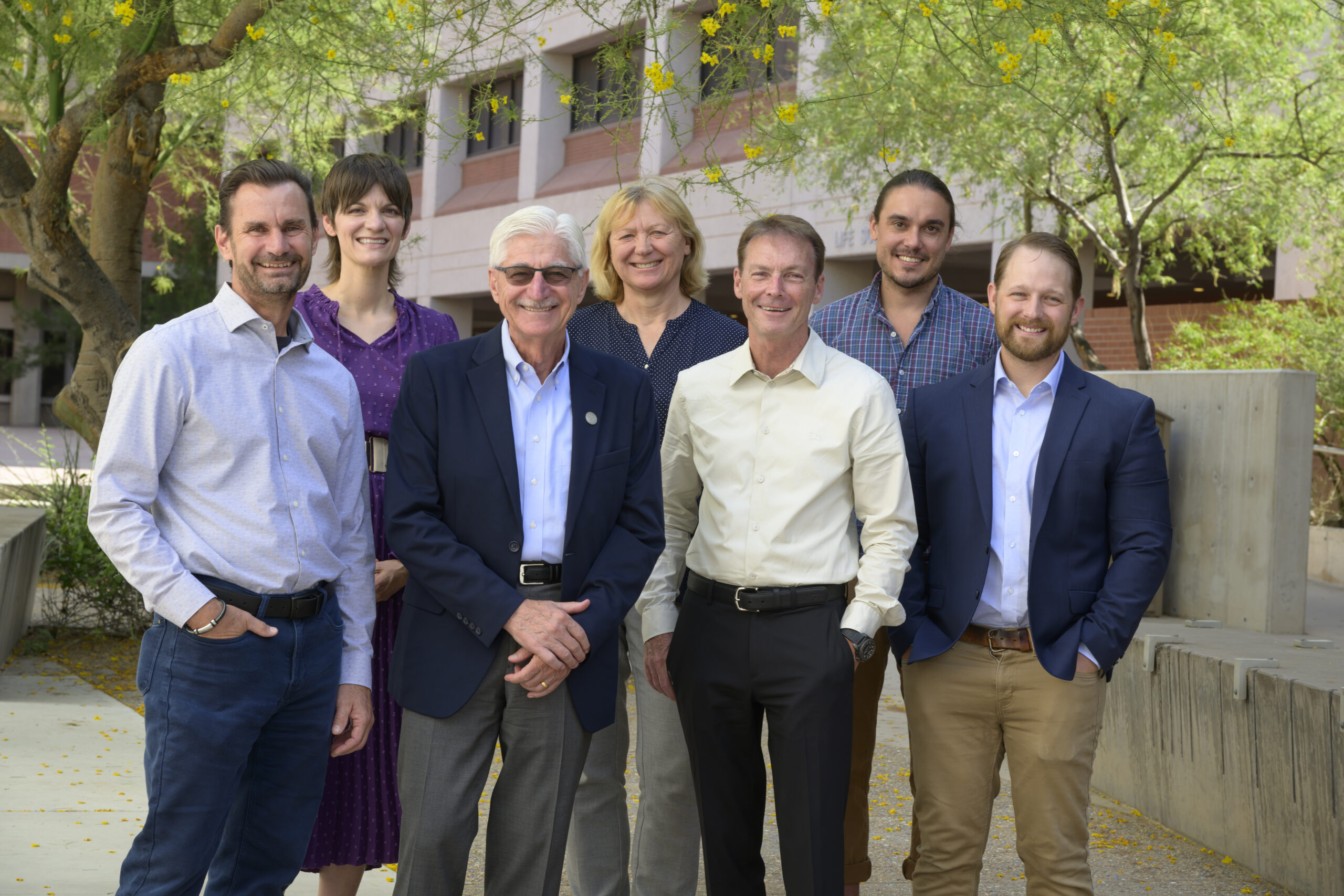 Group photo of seven members of a research team standing outdoors in front of a building with trees in the background. From left to right: Todd Vanderah, Amanda Wunsch, Frank Porreca, Edita Navratilova, Don Kyle, Justin LaVigne, and Craig Werner. They are smiling and dressed in professional attire. Photo by Kris Hanning, U of A Office of Research and Partnerships
